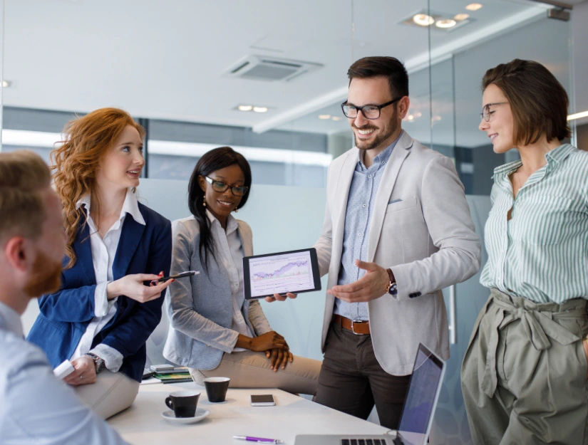 A professional group meeting, with a man presenting data on a tablet to colleagues in a modern office setting.