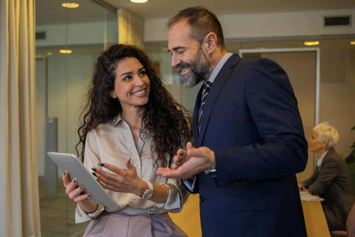 Two people in business attire having a conversation. One is holding a tablet. Another person is blurred in the background working at a desk.