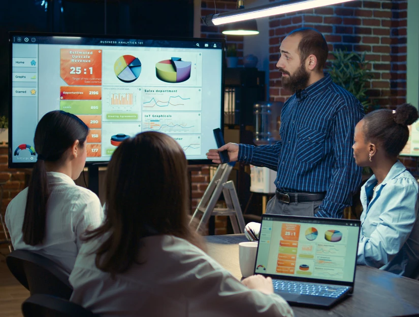 A group meeting with one person presenting business analytics data on a screen, while others observe and discuss with laptops open.