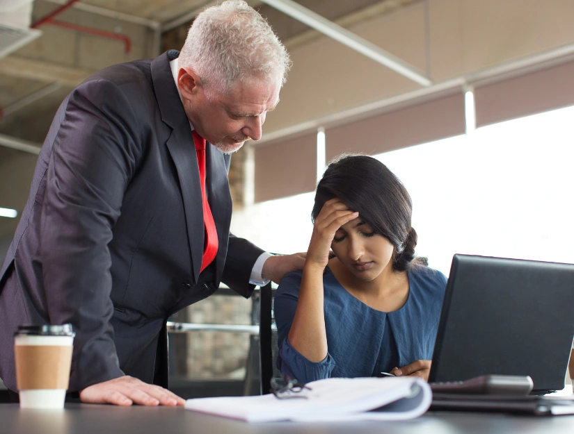 A person in a business suit leans over a seated colleague, discussing work at a desk with documents and a coffee cup nearby.
