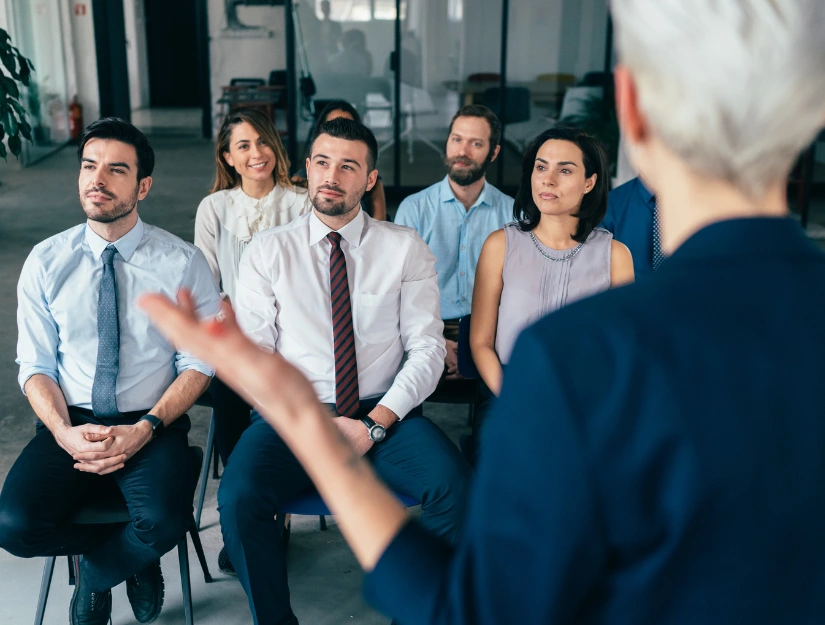 A group of professional individuals seated in a modern office, intently listening to a speaker gesturing in front of them.