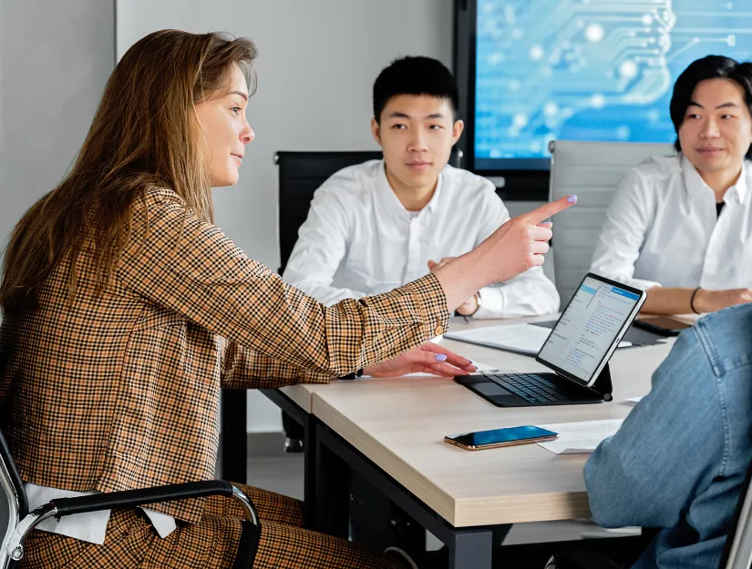 A businesswoman in a checked jacket gestures while discussing a presentation on a tablet with colleagues in a modern meeting room.