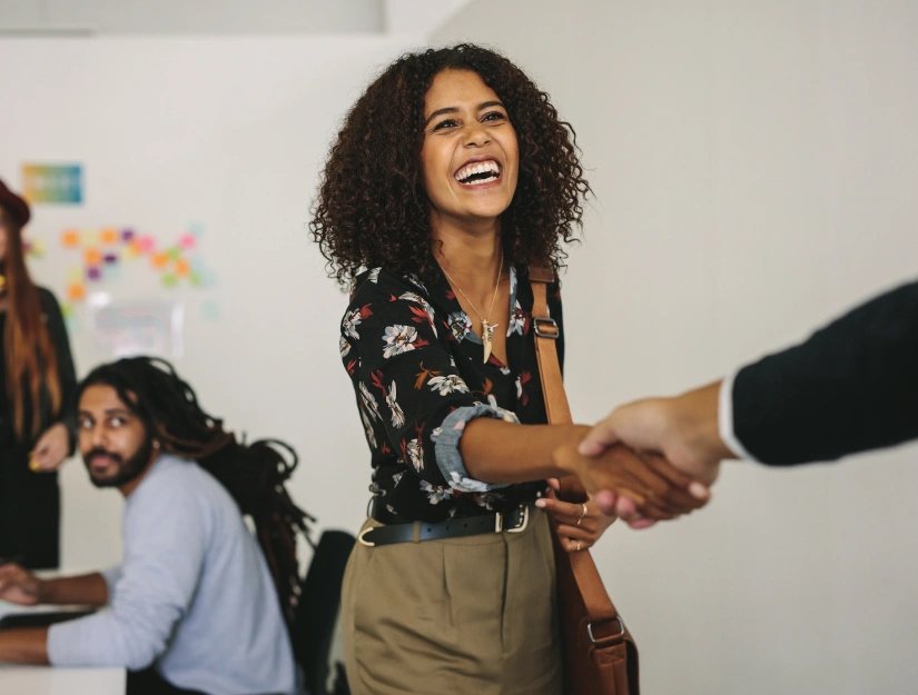 A woman with curly hair shakes hands in a modern office, while colleagues collaborate in the background. Colorful notes are on the wall.