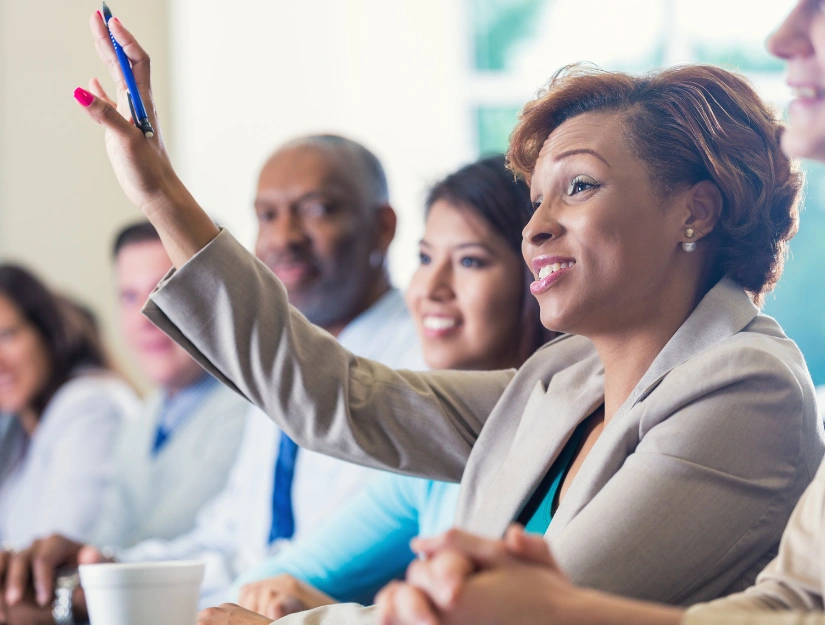 A group of professionals seated at a conference table, with one woman raising her hand to speak.