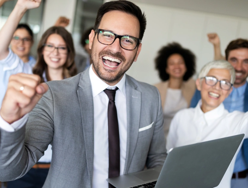 A group of business professionals celebrates success in a modern office space, with raised fists and a laptop in the foreground.