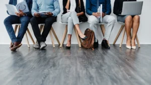 A diverse group of individuals in smart casual attire waits on chairs, engaged with devices and materials in a modern indoor setting.