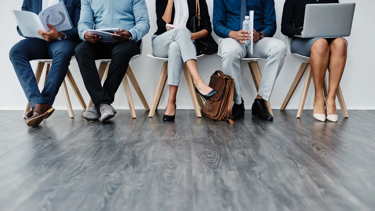 A diverse group of individuals in smart casual attire waits on chairs, engaged with devices and materials in a modern indoor setting.