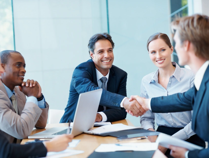 A group of business professionals engage in a handshake at a conference table, with a laptop and documents visible in the meeting room.