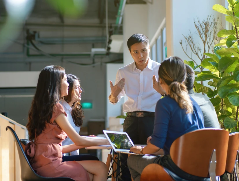 A group of five professionals engaged in a discussion in a bright, modern workspace, with plants in the background.