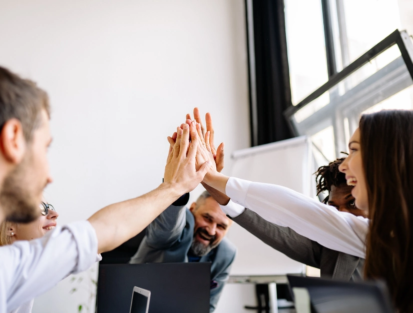 A diverse group of people joyfully joining hands in a high-five, celebrating teamwork in a bright, modern office setting.