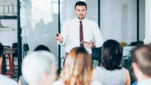A professional speaker presents to an audience in a modern office setting, engaging with listeners and emphasizing key points.