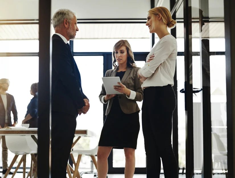 A group of professionals engage in a discussion in a modern office setting, with one person holding a tablet