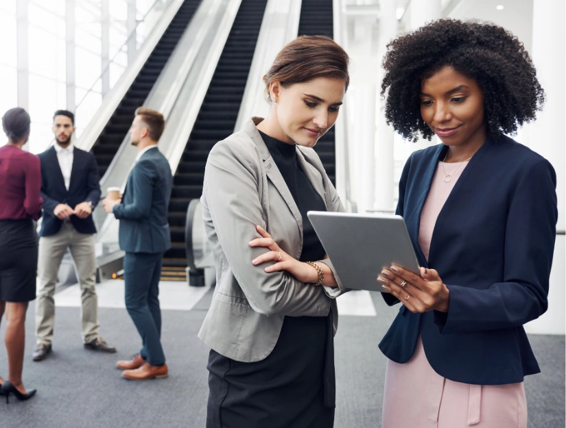 Two women in professional attire discuss information on a tablet while colleagues converse in the background near escalators