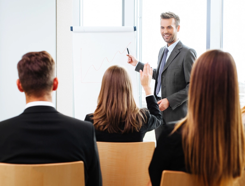 A businessman presents data on a flip chart to an audience in a modern conference room, with attendees engaged in discussion.