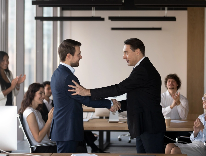Two men in suits shake hands in a modern office while colleagues applaud, celebrating a successful business agreement or achievement