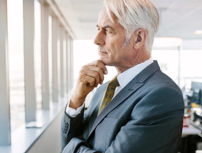 A thoughtful businessman in a suit stands by a large window, contemplating, with office workstations visible in the background.
