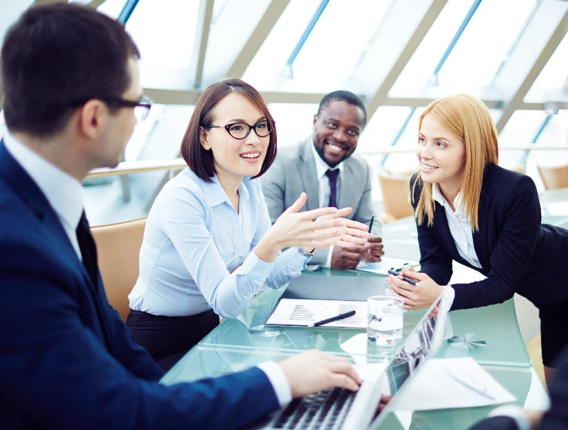 A group of professionals engaged in a discussion around a glass conference table with documents and a laptop in a bright office setting.