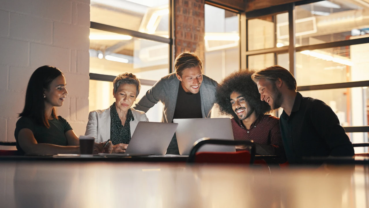 A team of professionals collaborates around laptops in a modern workspace, engaged in discussion and brainstorming ideas.