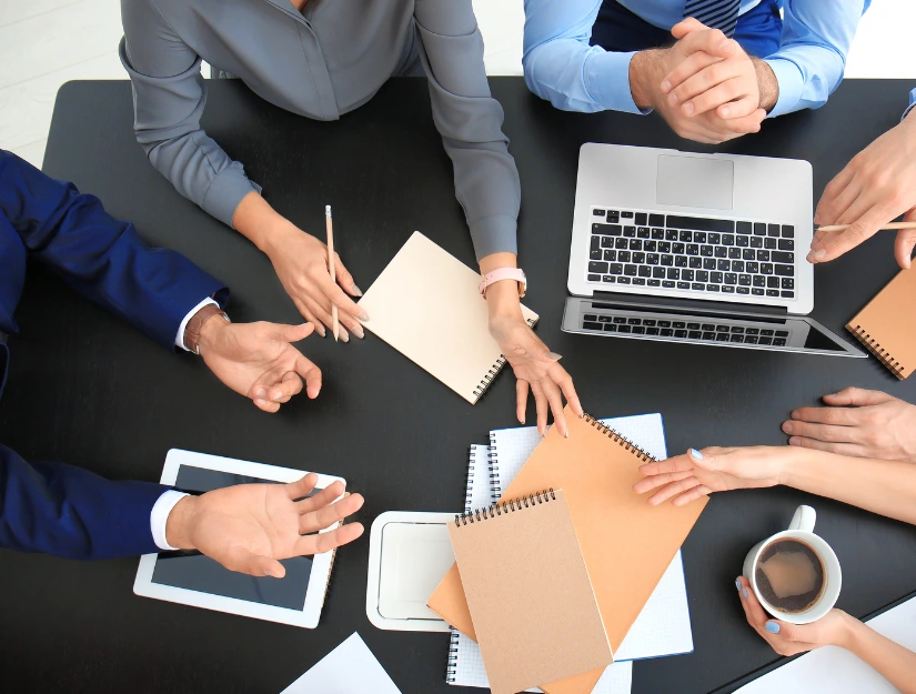 Aerial view of a collaborative meeting, featuring hands gesturing, notebooks, a laptop, and a coffee cup on a black table.