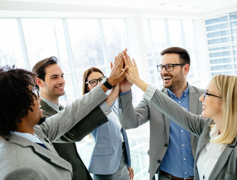 A group of business professionals in suits enthusiastically high-fiving each other in a bright, modern office space.