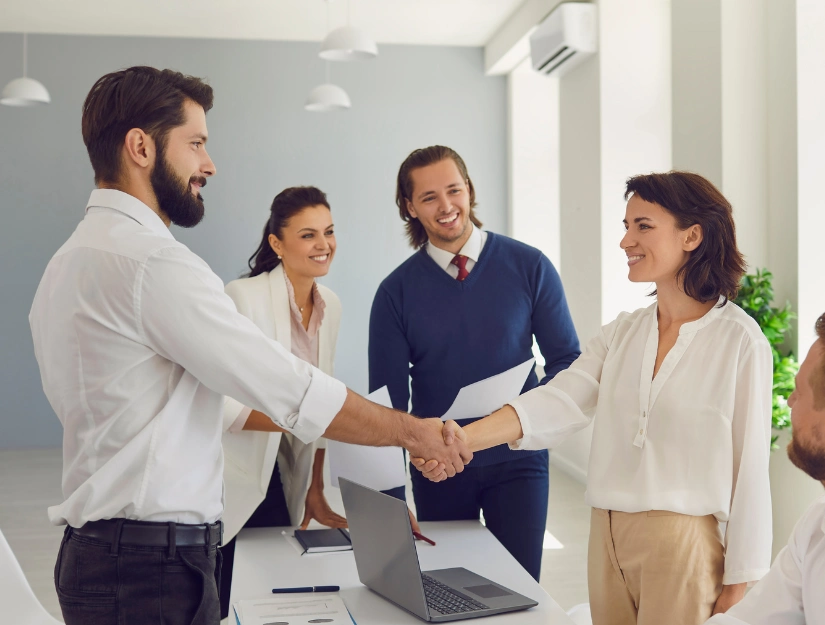 A group of professionals in a bright office space engaging in a handshake during a business meeting, with a laptop on the table.