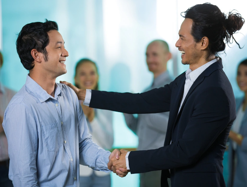 Two men engage in a handshake in a professional setting, with a group of blurred individuals in the background.