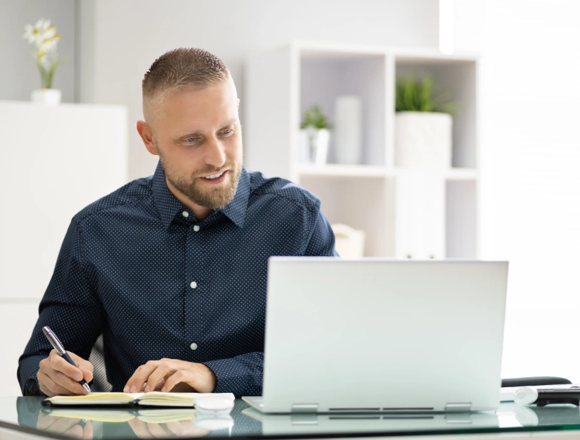 A person in a blue polka-dot shirt writes notes while sitting at a desk with a laptop, surrounded by indoor plants.