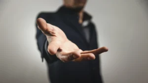 A man in a black suit extends a hand forward, suggesting an invitation or greeting against a neutral background.