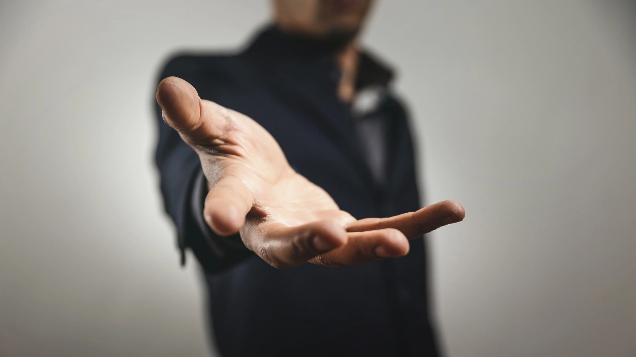 A man in a black suit extends a hand forward, suggesting an invitation or greeting against a neutral background.