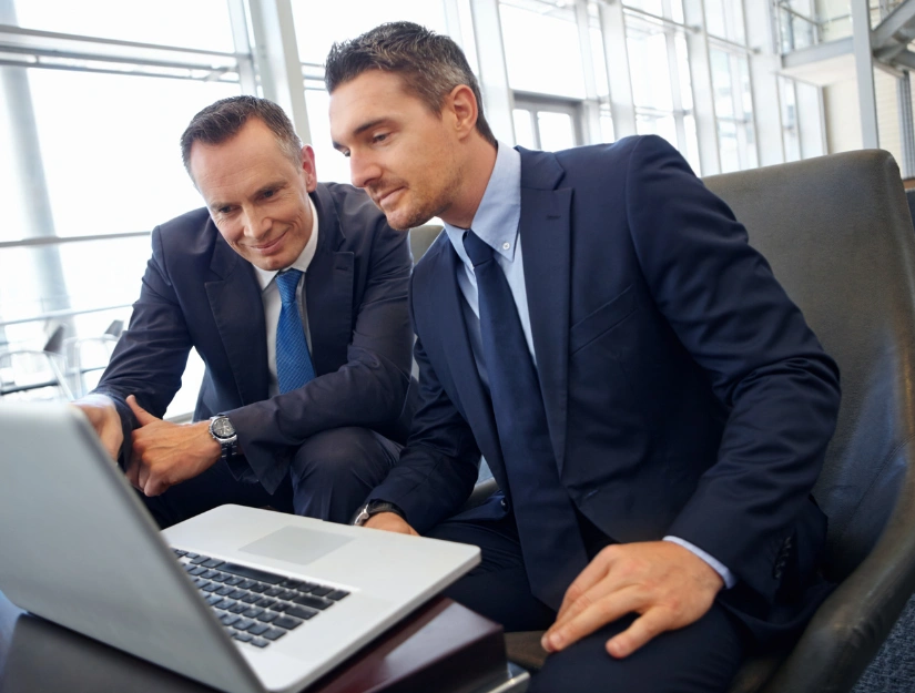 Two businessmen in suits sit in an office setting, looking at a laptop screen and discussing work.