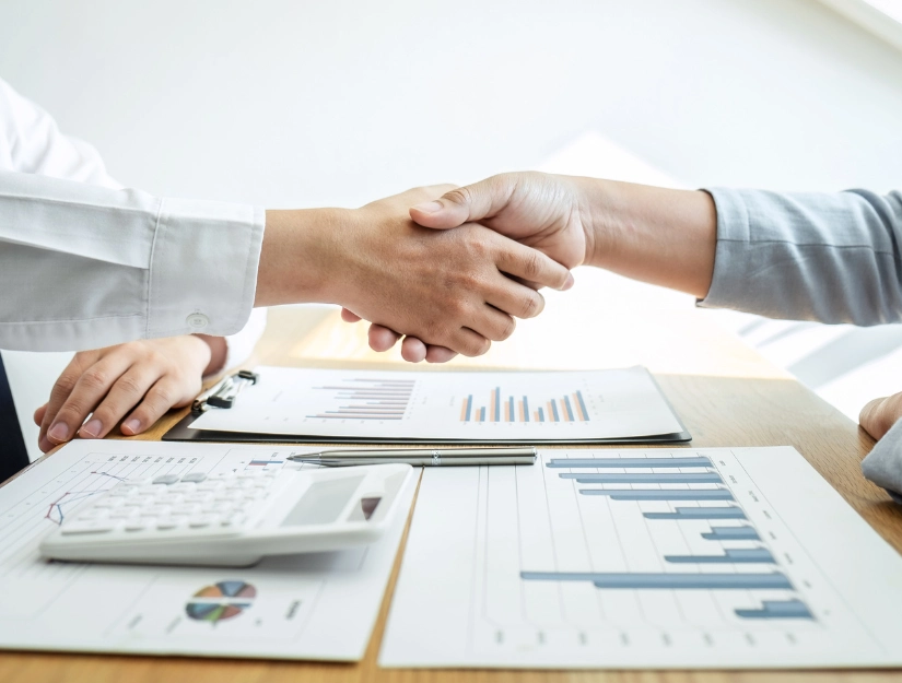 Two professionals shake hands over a table with financial reports and a calculator.