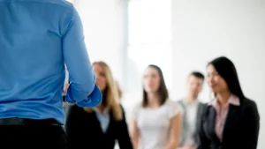 A person in a blue shirt stands in front of a group of professionals, who appear engaged in a discussion or presentation.