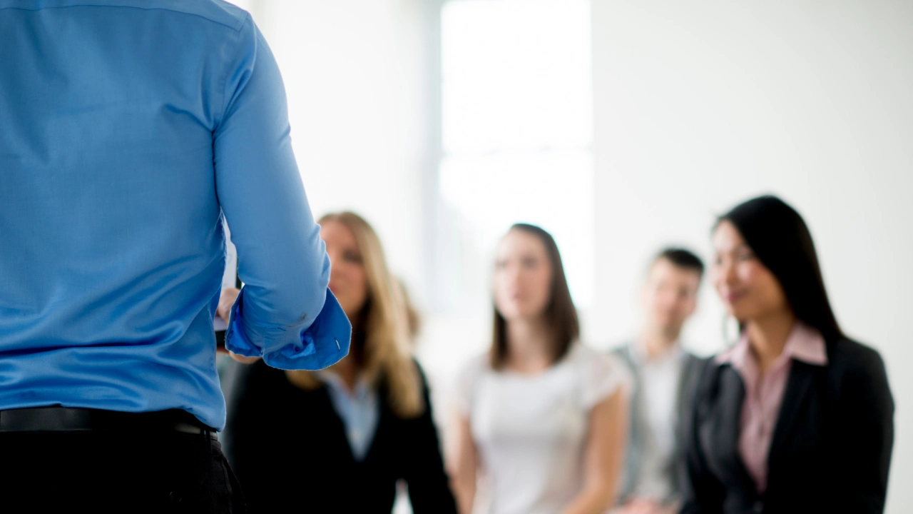 A person in a blue shirt stands in front of a group of professionals, who appear engaged in a discussion or presentation.