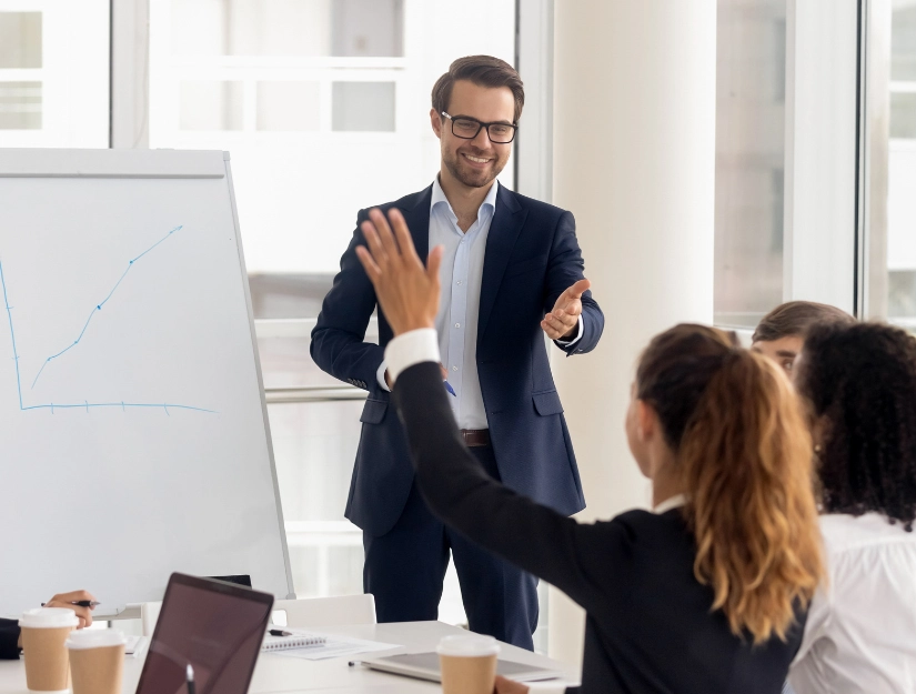 A business professional presents a graph to an engaged audience in a modern meeting room, with participants actively discussing.