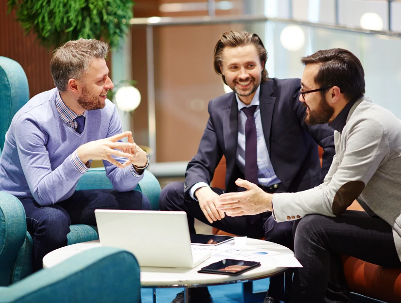 Three professionals engage in a lively discussion around a laptop in a modern, stylish seating area with vibrant upholstery.
