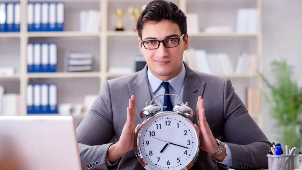 A businessman in a suit holds a large alarm clock, emphasizing the concept of time management in a modern office setting.
