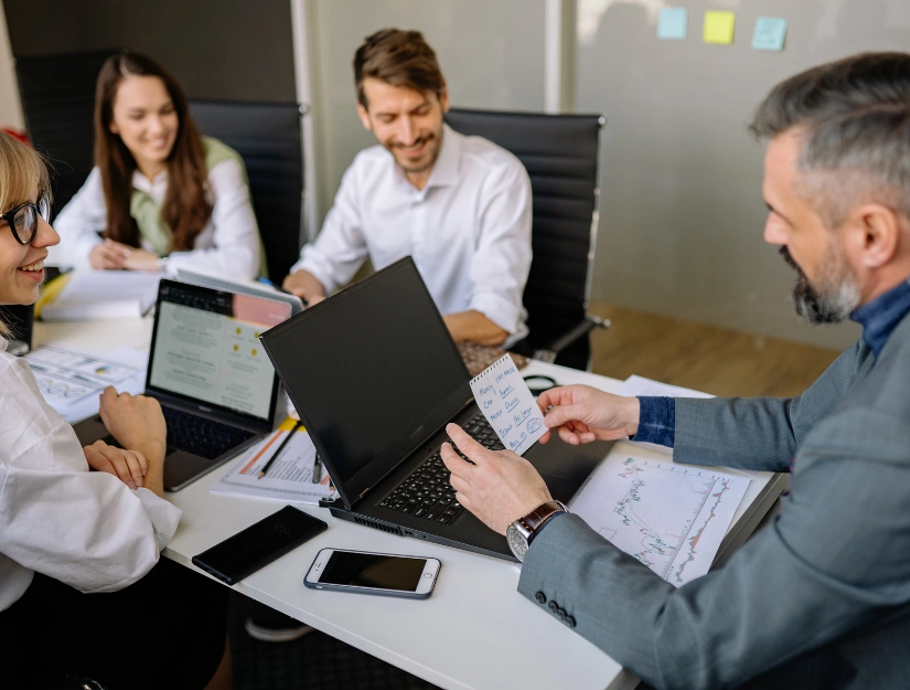 A team of professionals collaborates around a table, discussing strategies with laptops and documents in a modern office setting.