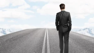 A man in a business suit walks away on a solitary road, surrounded by mountains under a bright blue sky with fluffy clouds.