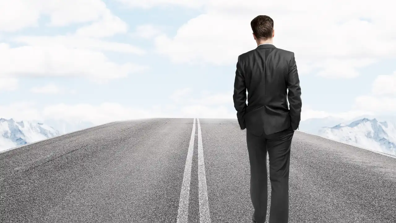 A man in a business suit walks away on a solitary road, surrounded by mountains under a bright blue sky with fluffy clouds.