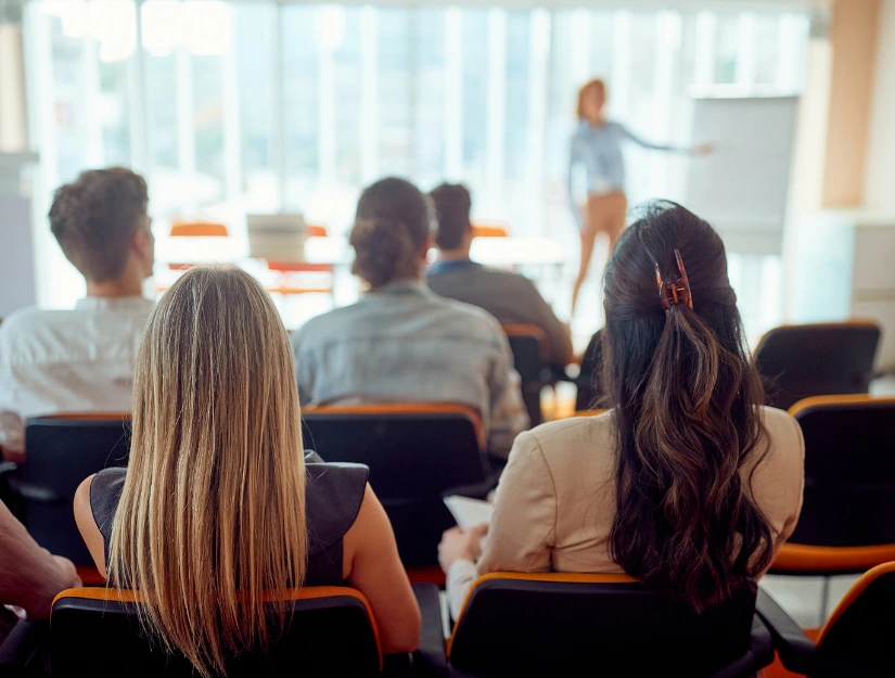 A group of people seated in a classroom listens to a presenter standing near a whiteboard in front of large windows.