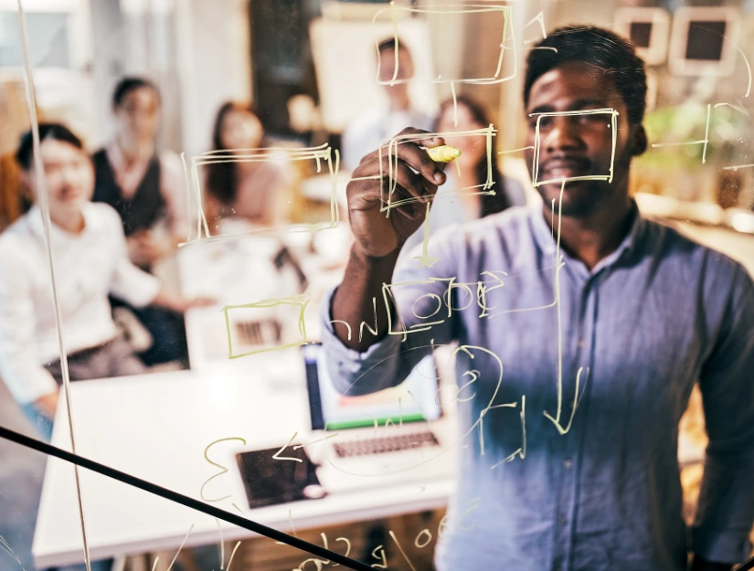 A person writes on a glass board with yellow markers, while a group of people observes in a modern office setting.