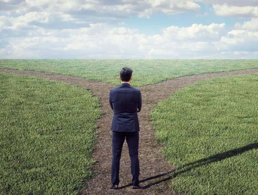 A man in a suit stands at a fork in a dirt path on a grassy field, facing away and contemplating which direction to take under a partly cloudy sky.